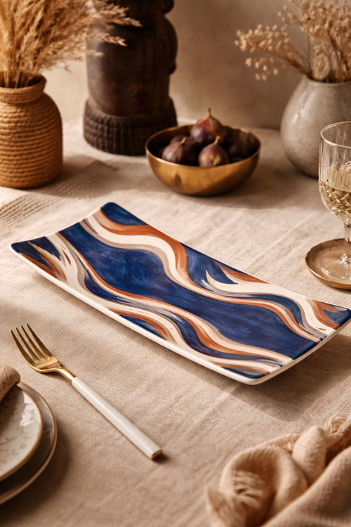 Mise en scène photographique d’une assiette rectangulaire Mbeya en céramique de designer, décorée d’un motif ondulé bleu profond, terracotta et sable, présentée sur une table élégante avec textile naturel, vaisselle raffinée et éléments décoratifs d’inspiration afro-luxe.