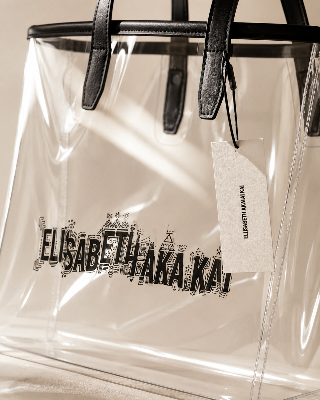 Decouvrez les finitions du sac cabas transparent Elisabeth Akaia Kai. Transparence, coutures precises, anses noires et logo graphique se revelent dans une photographie de maroquinerie haut de gamme.