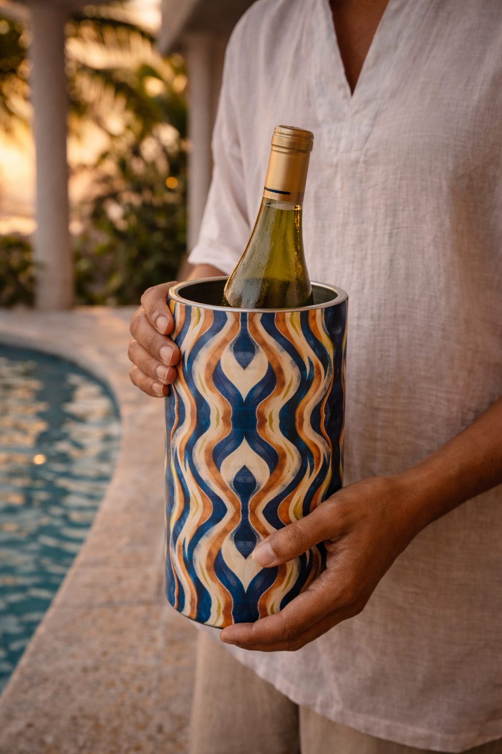 Une personne porte délicatement un rafraîchisseur de vin Mbeya en acier inoxydable contenant une bouteille de champagne, scène en extérieur en fin de journée, ambiance afro-luxe élégante au bord de la piscine à Libreville.