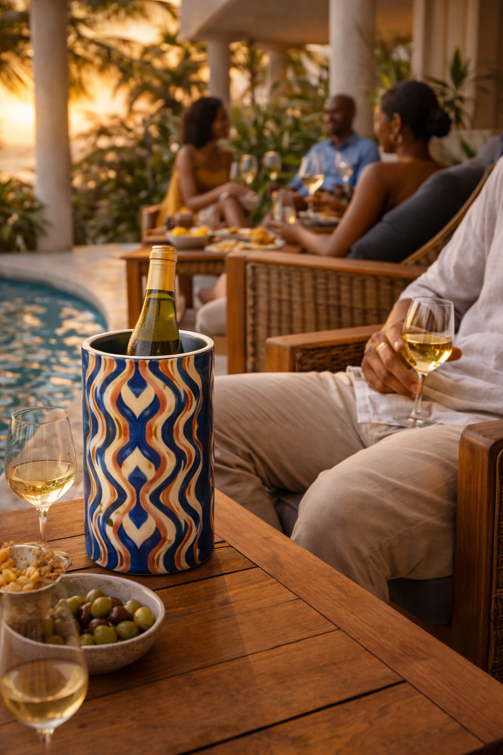 Rafraîchisseur de vin Mbeya en acier inoxydable posé sur une table basse en bois à côté d’un fauteuil d’extérieur tressé, bouteille de vin blanc à l’intérieur, groupe d’amis prenant l’apéritif au bord de la piscine en fin de journée, ambiance afro-luxe élégante et paisible.