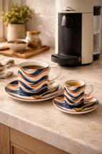 Tasse à café et tasse expresso design Mbeyla avec leurs sous-tasses assorties, posées sur un plan de travail de cuisine aux côtés d’une machine à café de style Vertuo, mise en scène élégante évoquant un art de vivre afro-luxe, le rituel du café et le design gabonais contemporain.