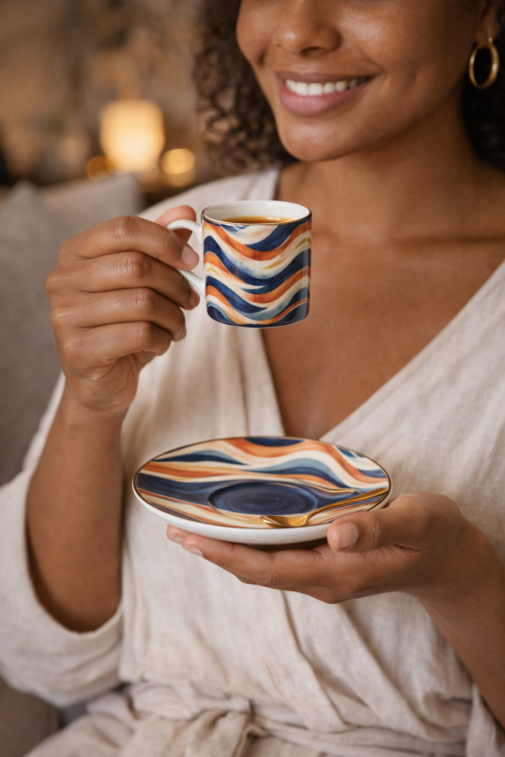 Femme souriante savourant un espresso dans une tasse design Mbeyla aux motifs ondulations solaires bleu, terracotta et sable, tenant la sous-tasse assortie dans un salon chaleureux, photographie d’objet en situation évoquant un art de vivre afro-luxe et un moment de calme intérieur.