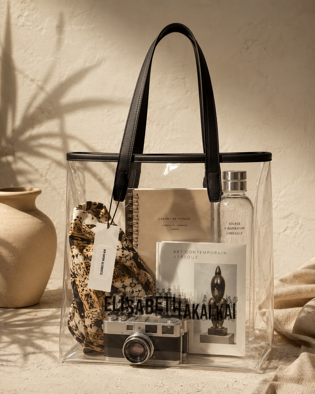 Le sac cabas transparent Elisabeth Akaia Kai accueille un carnet de voyage, un foulard artistique, un livre d'art et un appareil photo argentique. Une creation pensee pour les voyageurs en quete d'elegance.