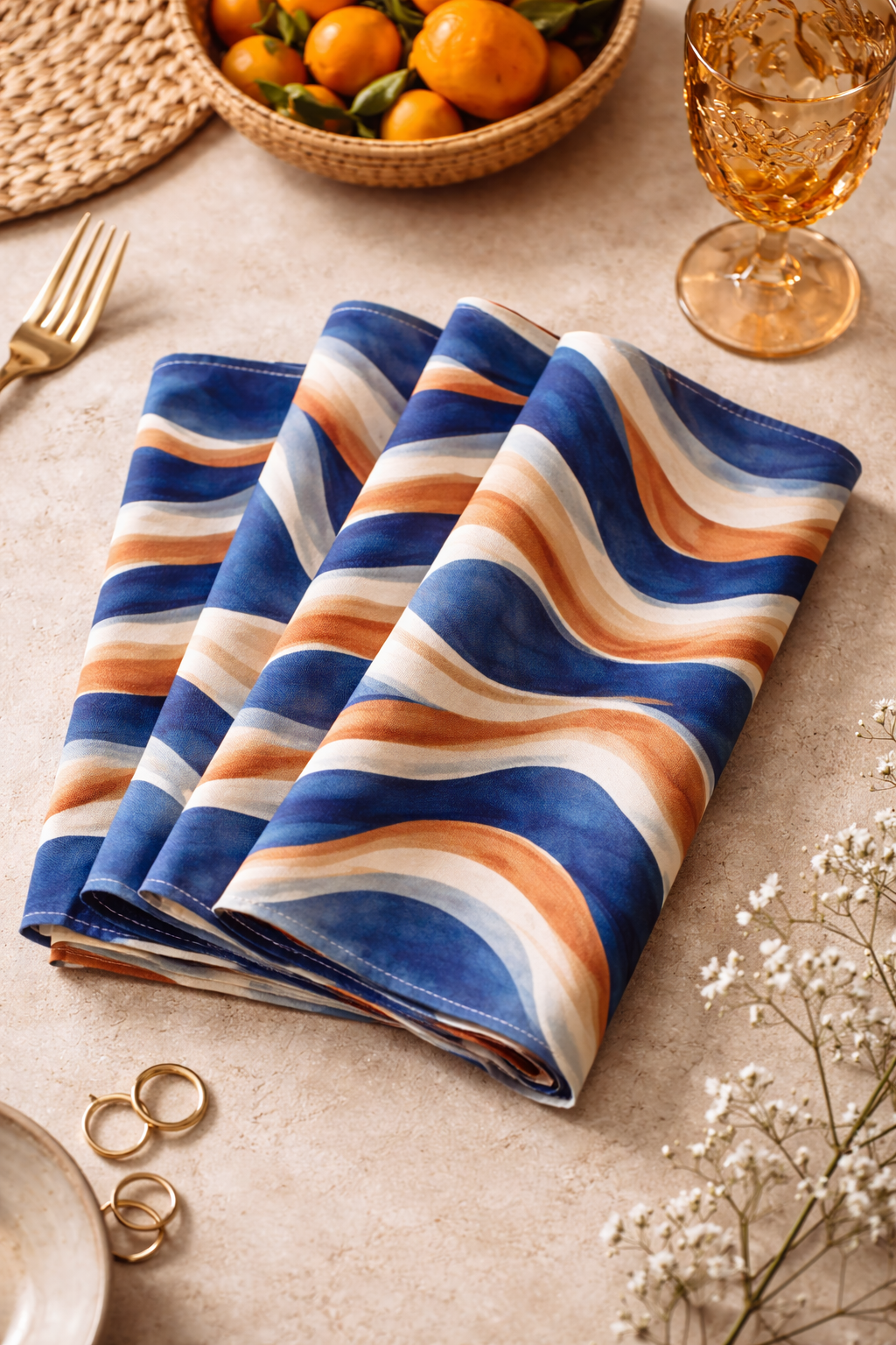 Photographie de mise en scène montrant plusieurs serviettes de table Mbyela 40 × 40 cm pliées, aux motifs ondulations solaires bleu profond, terracotta et sable, présentées sur une surface claire avec accessoires raffinés, illustrant un art de la table design et luxe contemporain.