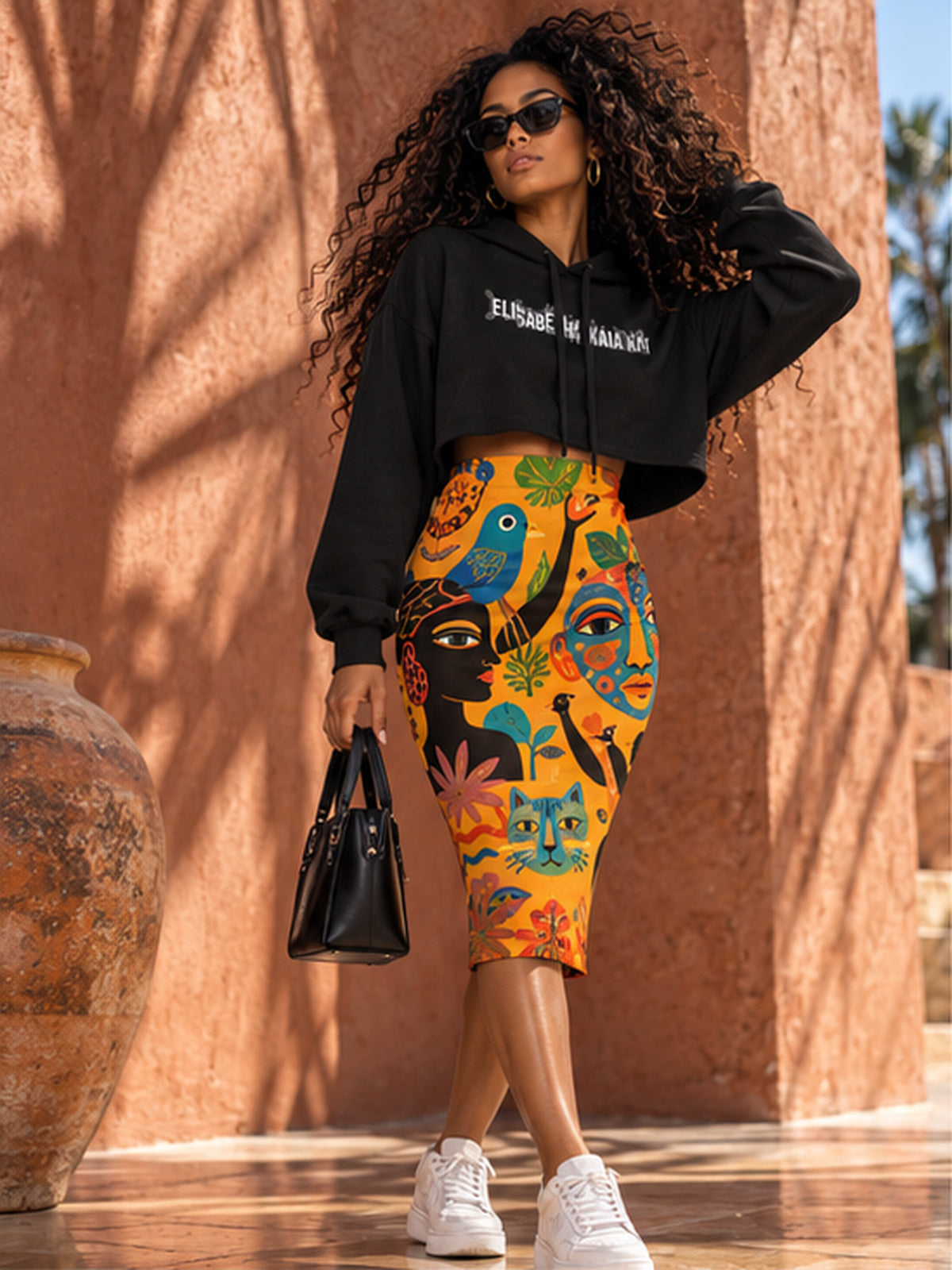 Mannequin portant le sweat court noir EAK avec une jupe crayon imprimée de visages et de motifs graphiques, dans l'univers afro-luxe de la Maison.