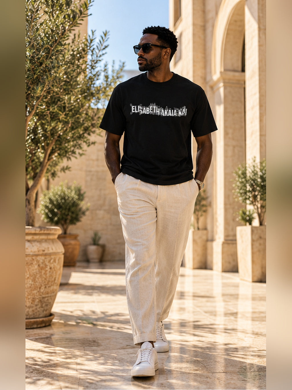 Mannequin homme marchant dans une cour méditerranéenne élégante, vêtu du t-shirt KAÏ noir à logo blanc et d'un pantalon en lin écru.