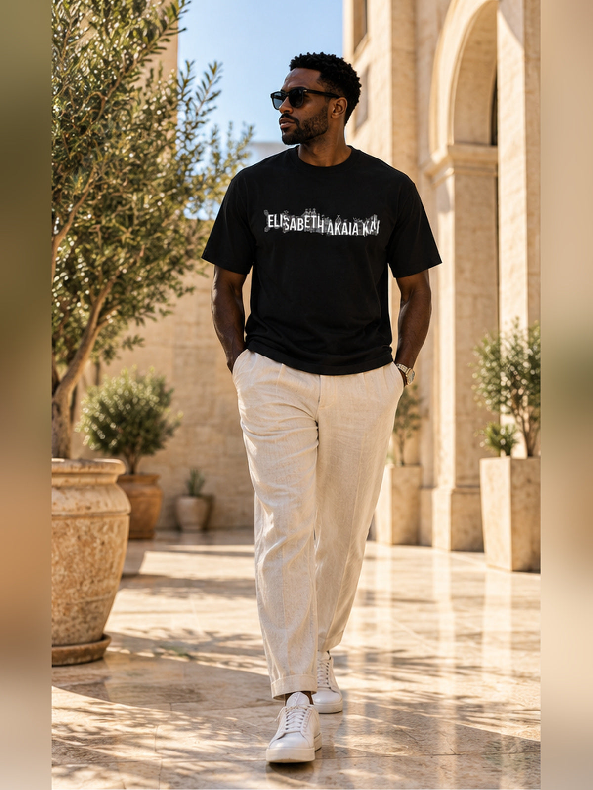 Mannequin homme marchant dans une cour méditerranéenne élégante, vêtu du t-shirt KAÏ noir à logo blanc et d'un pantalon en lin écru.