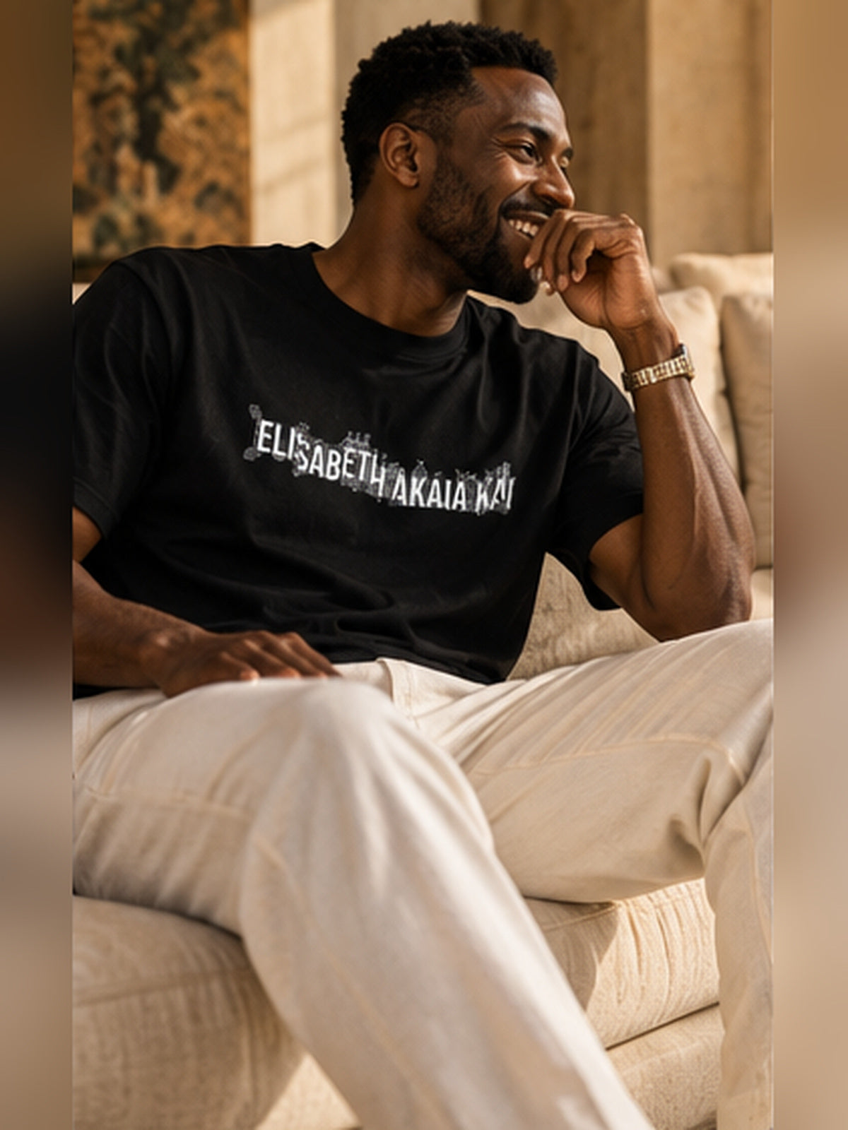 Un homme souriant porte le t-shirt KAÏ noir à logo blanc avec un pantalon écru, assis dans un salon élégant baigné de lumière naturelle.