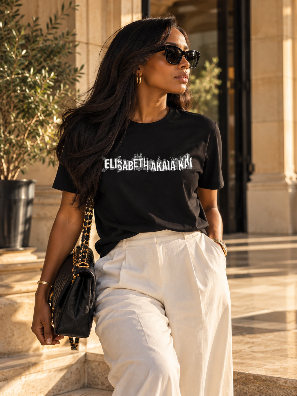 Femme portant le t-shirt KAÏ noir à signature blanche, un pantalon ivoire et un sac noir devant des colonnes en pierre baignées de soleil.