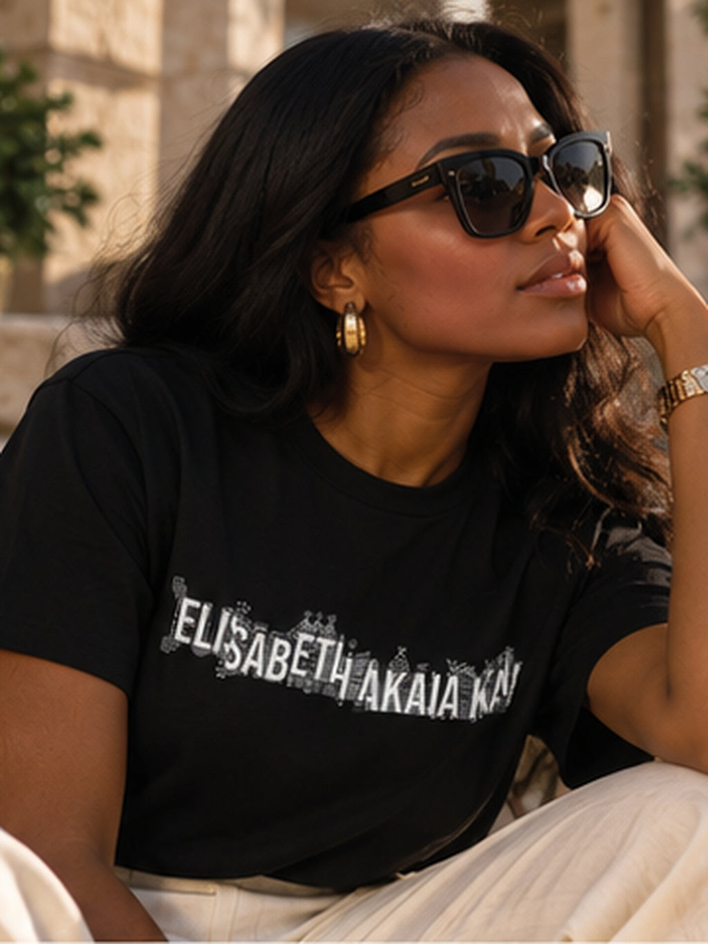 Portrait d'une femme assise portant le t-shirt KAÏ noir de créateur, associé à un pantalon écru, des lunettes et des bijoux dorés.